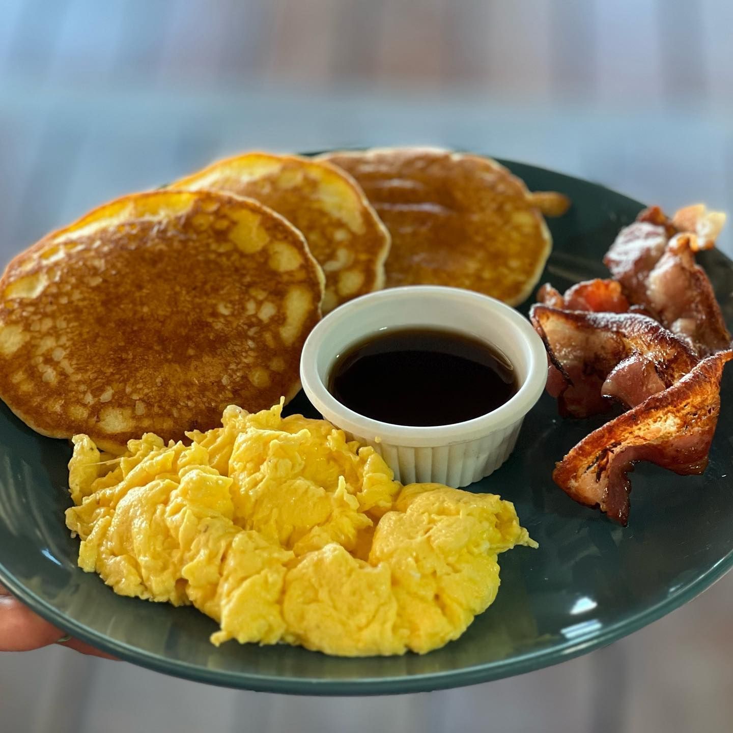 A person is holding a plate of pancakes scrambled eggs bacon and syrup.
