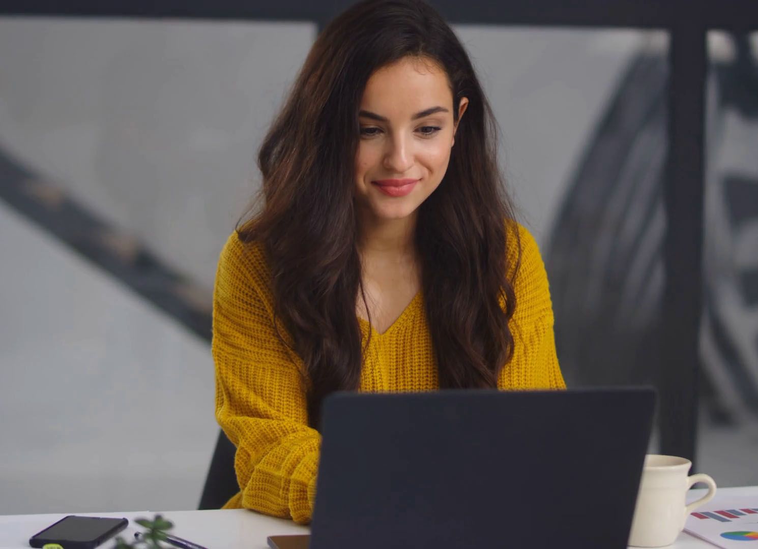 Una mujer con un suéter amarillo está sentada en un escritorio usando una computadora portátil.