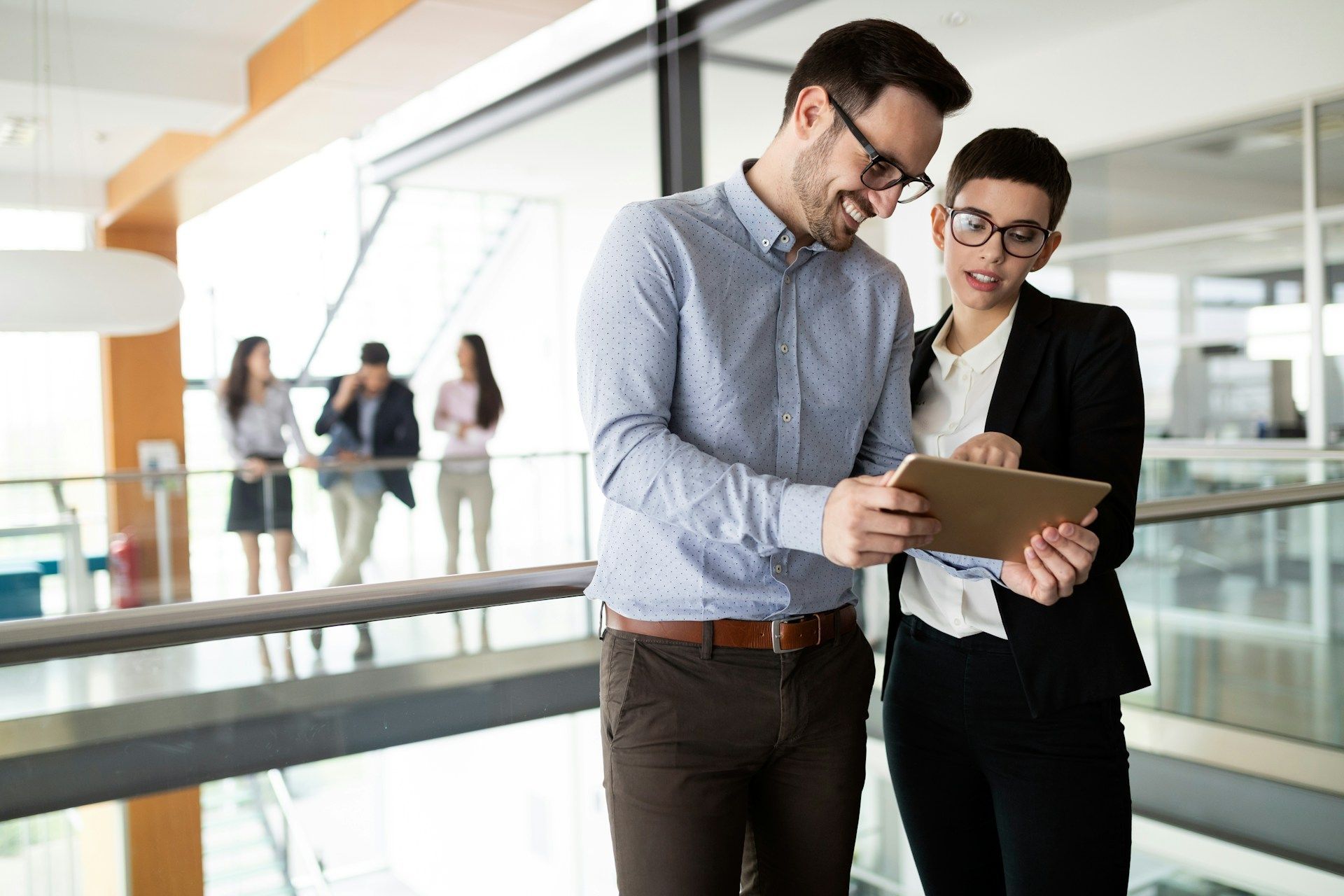 Two business professionals review a tablet in a modern office setting; others work in the background.