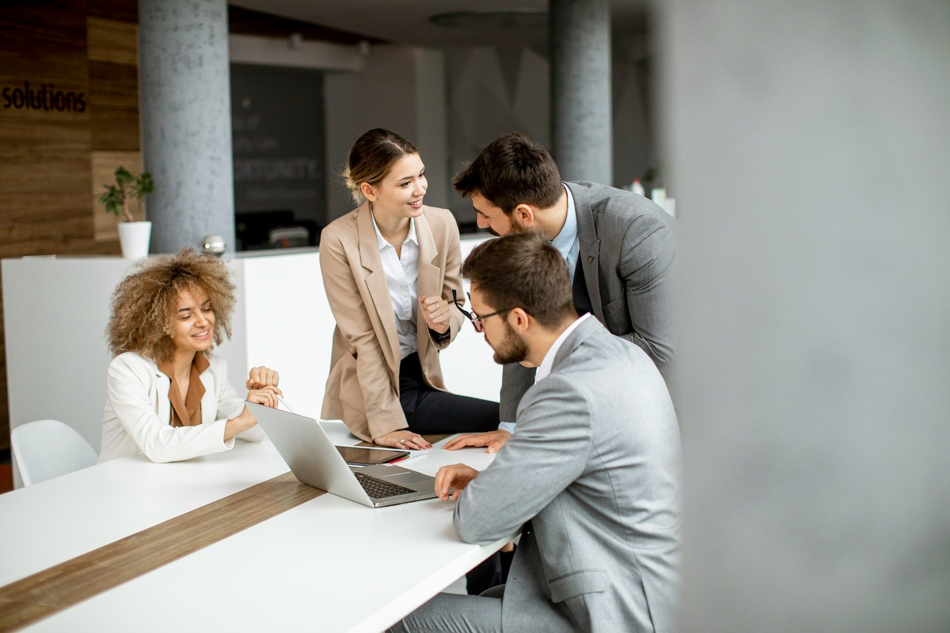 Four colleagues in suits gather around a laptop in a modern office, collaborating on a project.