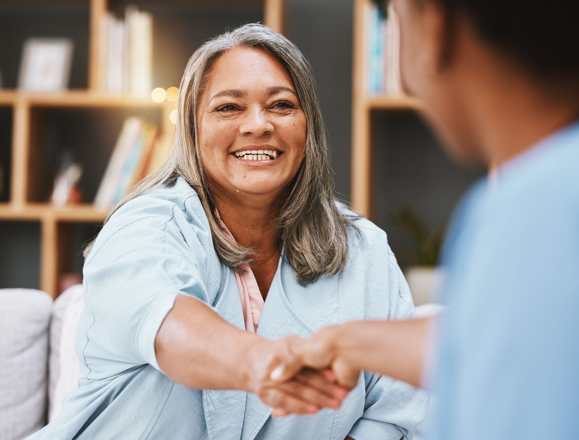 Smiling older woman with gray hair shakes hands with another person in a warm, indoor setting.