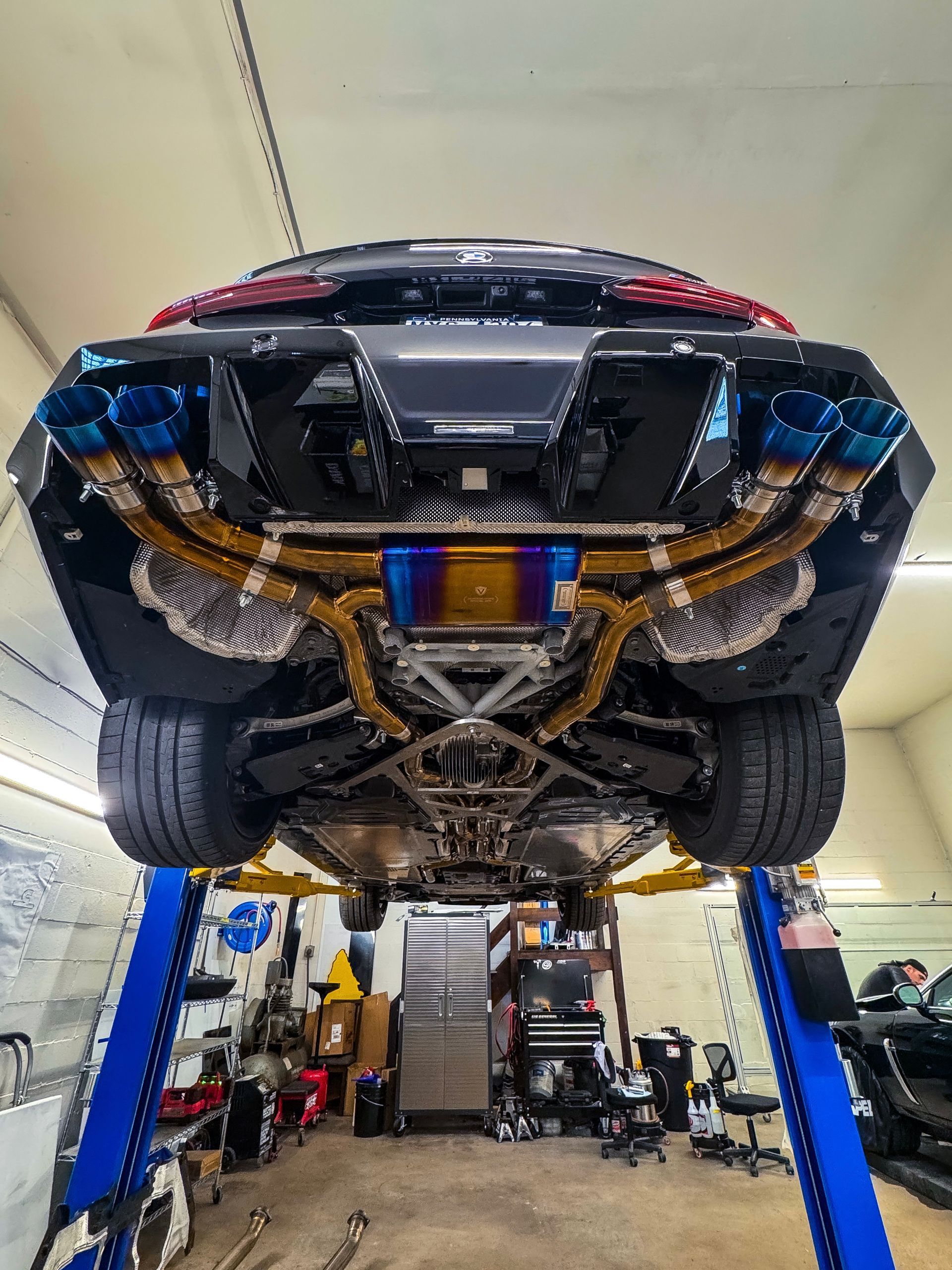 Black sports car elevated on a lift, revealing custom gold exhaust system with blue tips, in a garage.