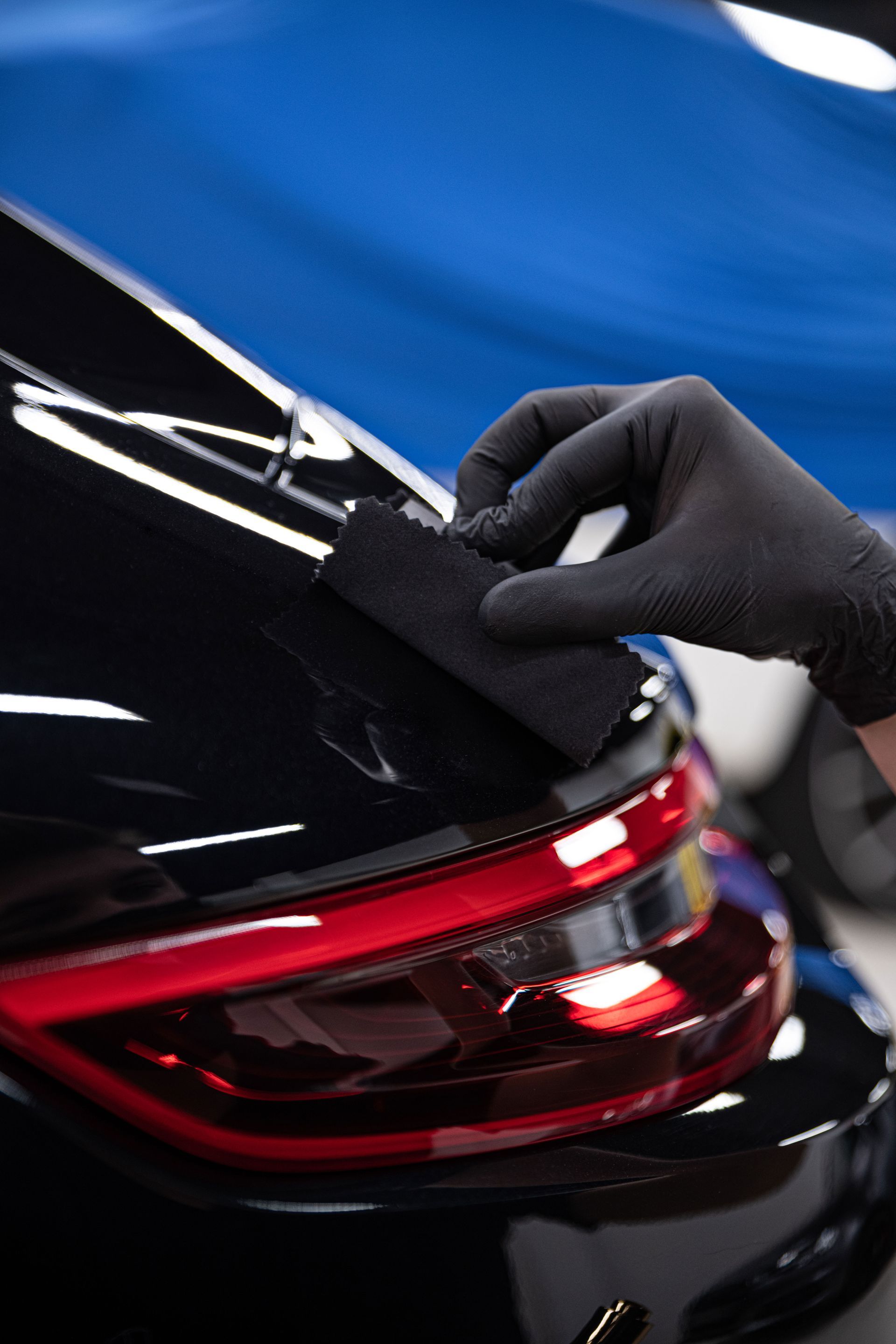 Gloved hand applying a coating to a black car's taillight with a black applicator in a workshop setting.