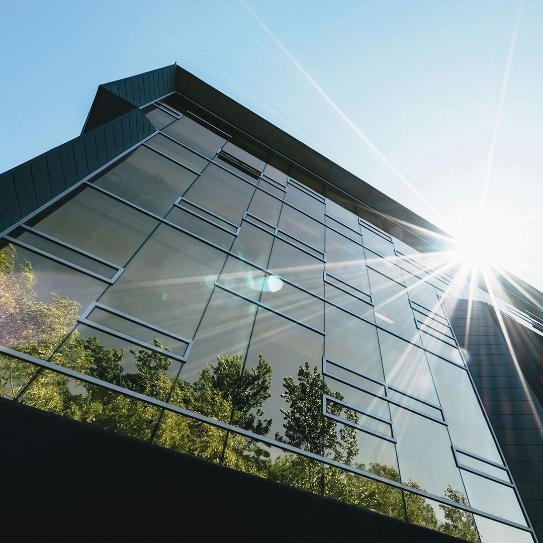Modern office building with reflective glass facade, bright sunlight, and green trees.