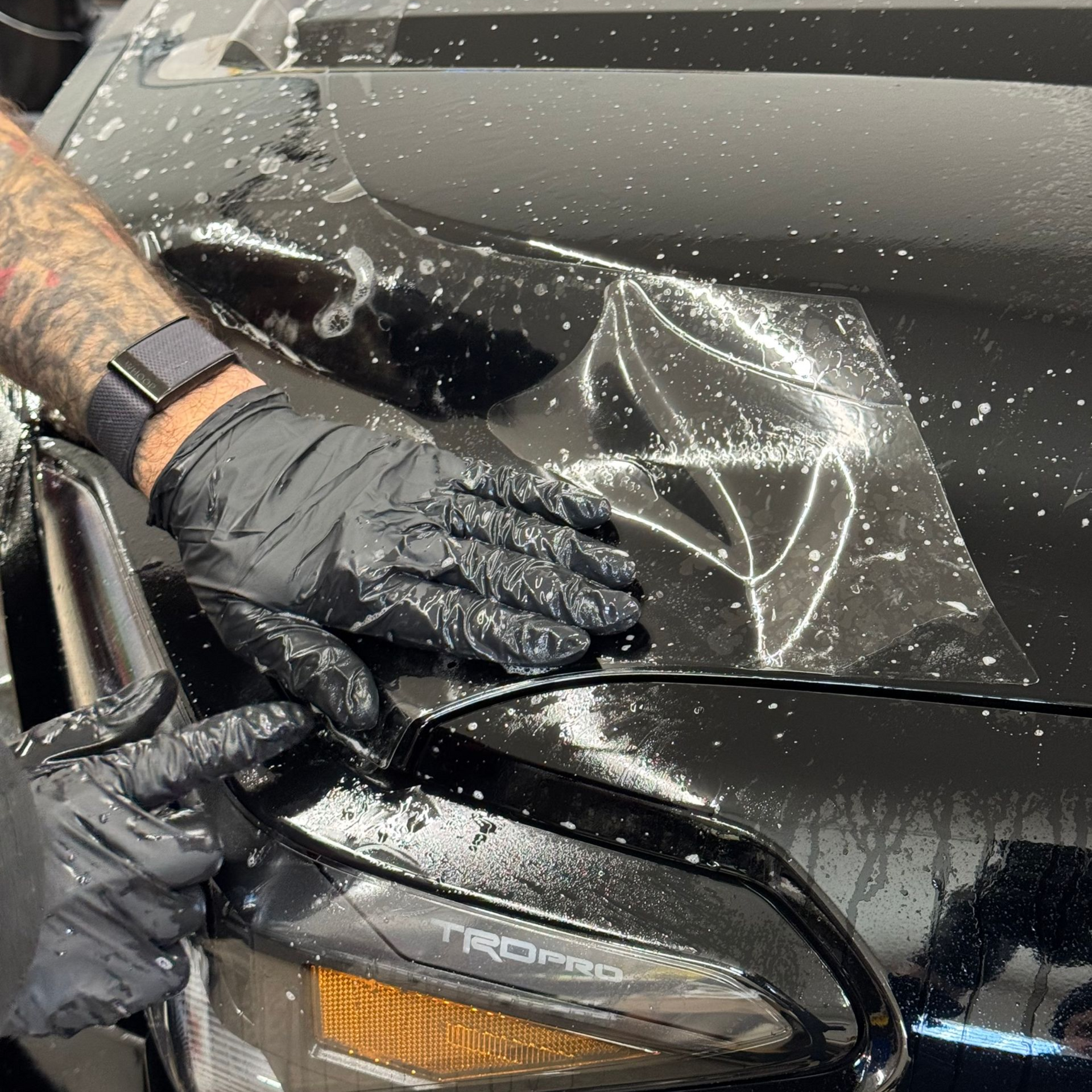 Person in black gloves applying film to a black car hood. Spray and light reflection visible.