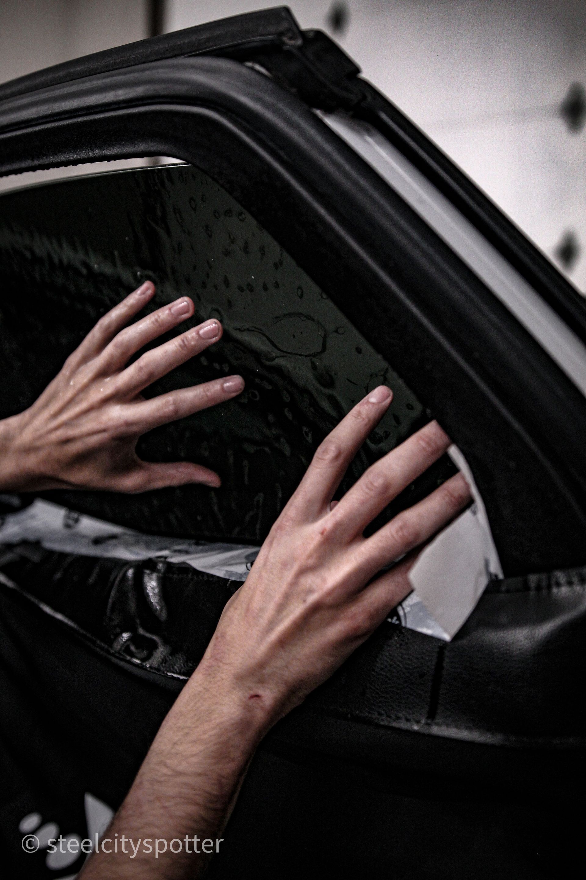 Person's hands smoothing window tint onto a car window; black vehicle, indoor setting.