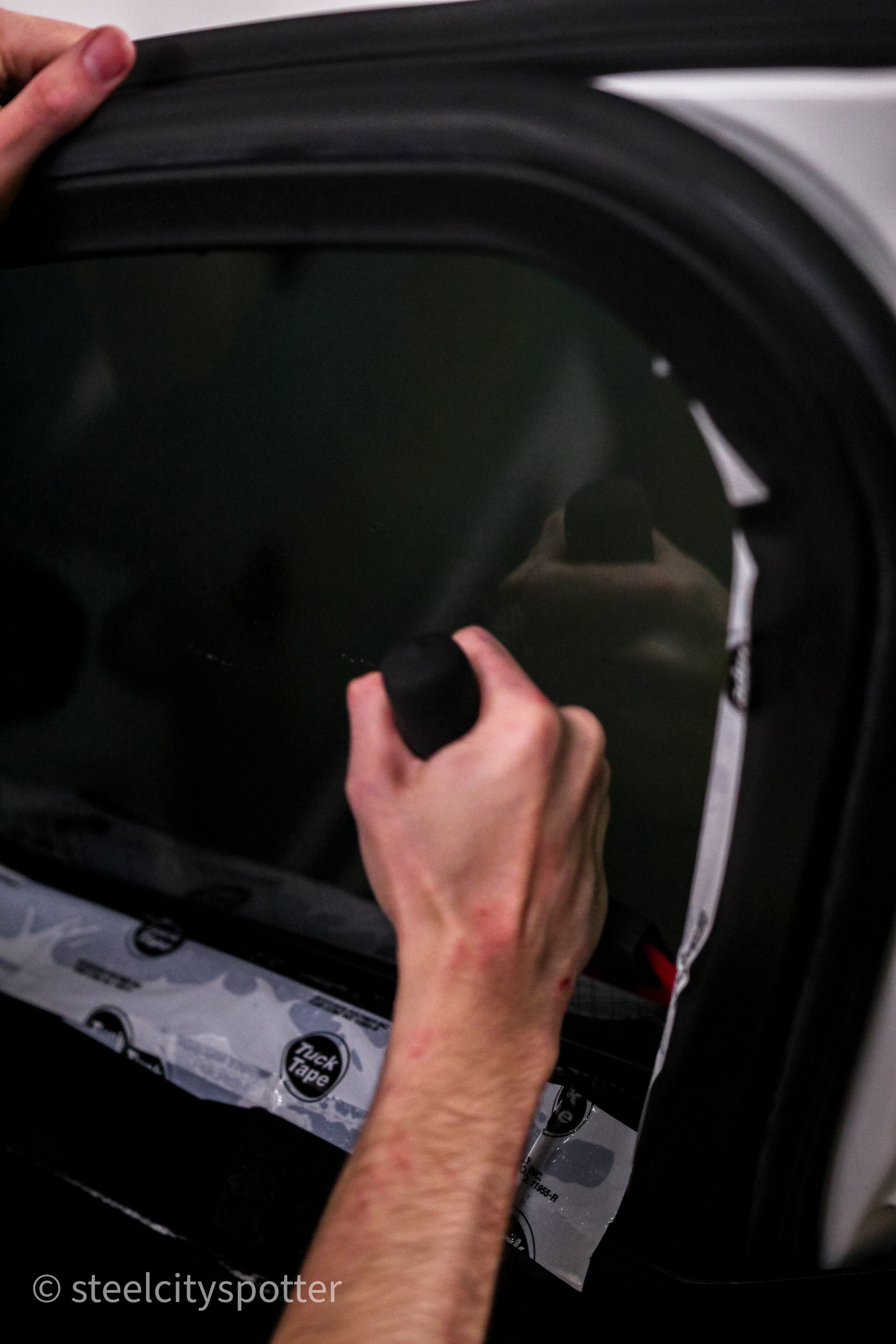 Hands applying tint film to a car window. Black film, white trim.
