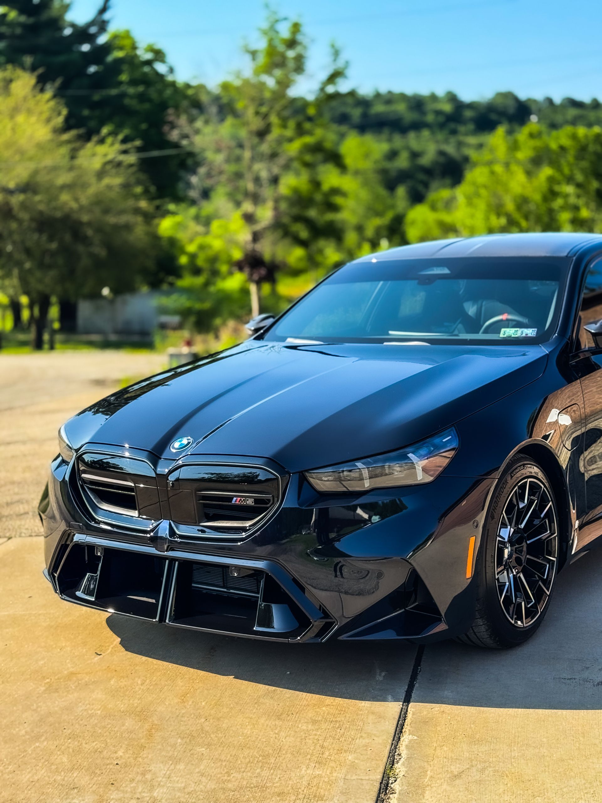 Black BMW sedan parked on a driveway with trees and hills in the background.