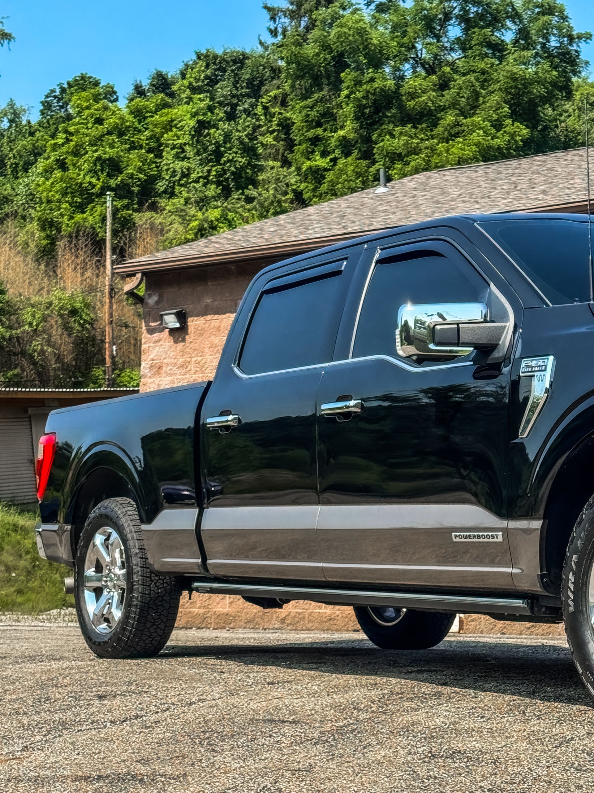 Black Ford pickup truck parked on gravel, tan trim, chrome accents, near a building and trees.
