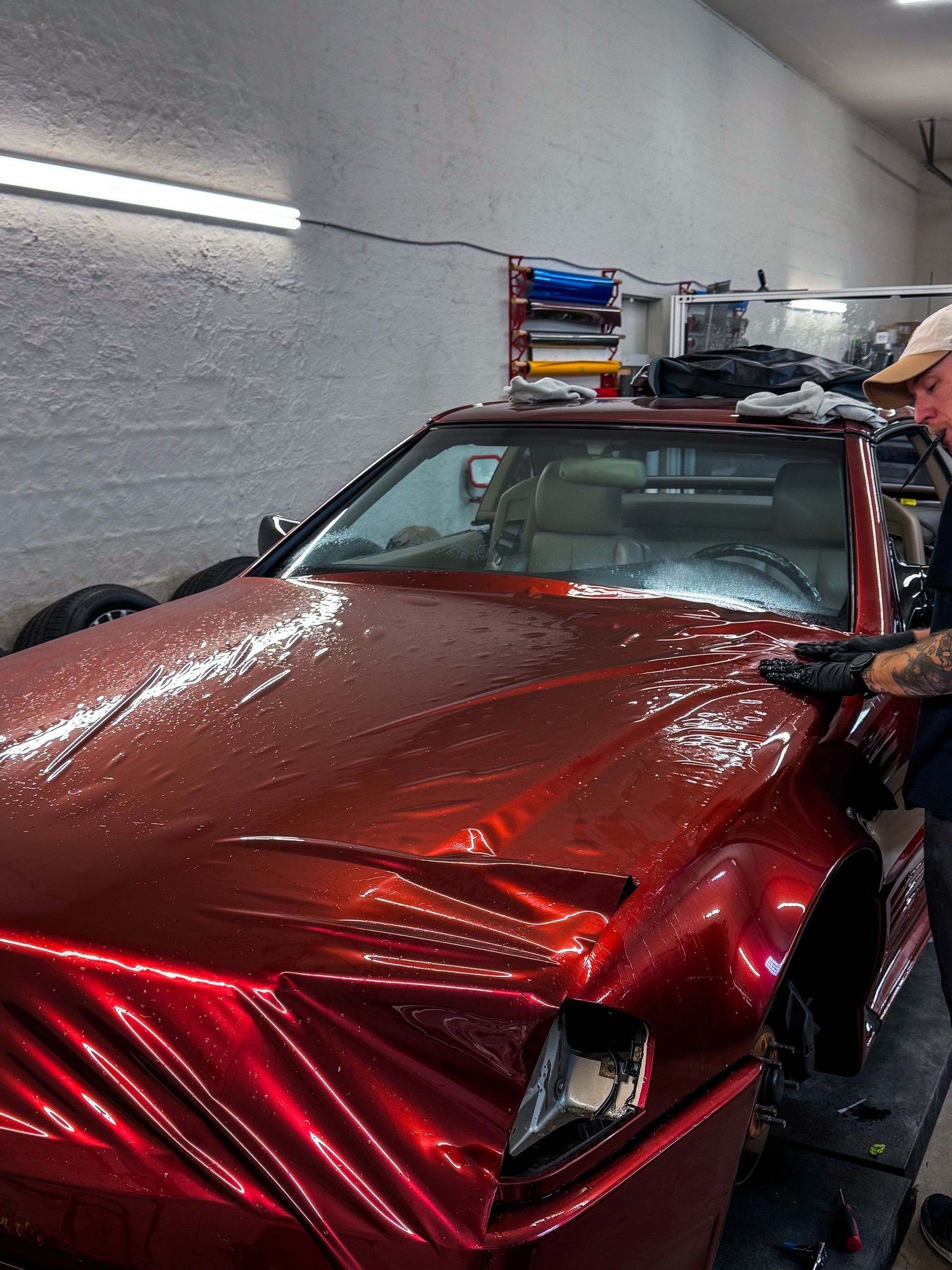 Red car being wrapped in shiny material by a person in a workshop.