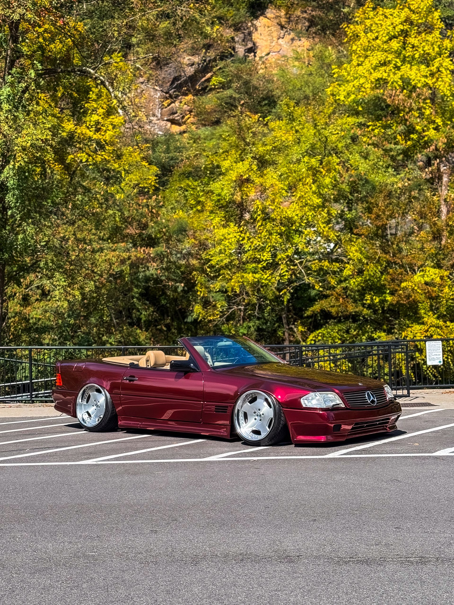 Burgundy convertible car parked with lowered suspension, against a backdrop of trees and a rock face.