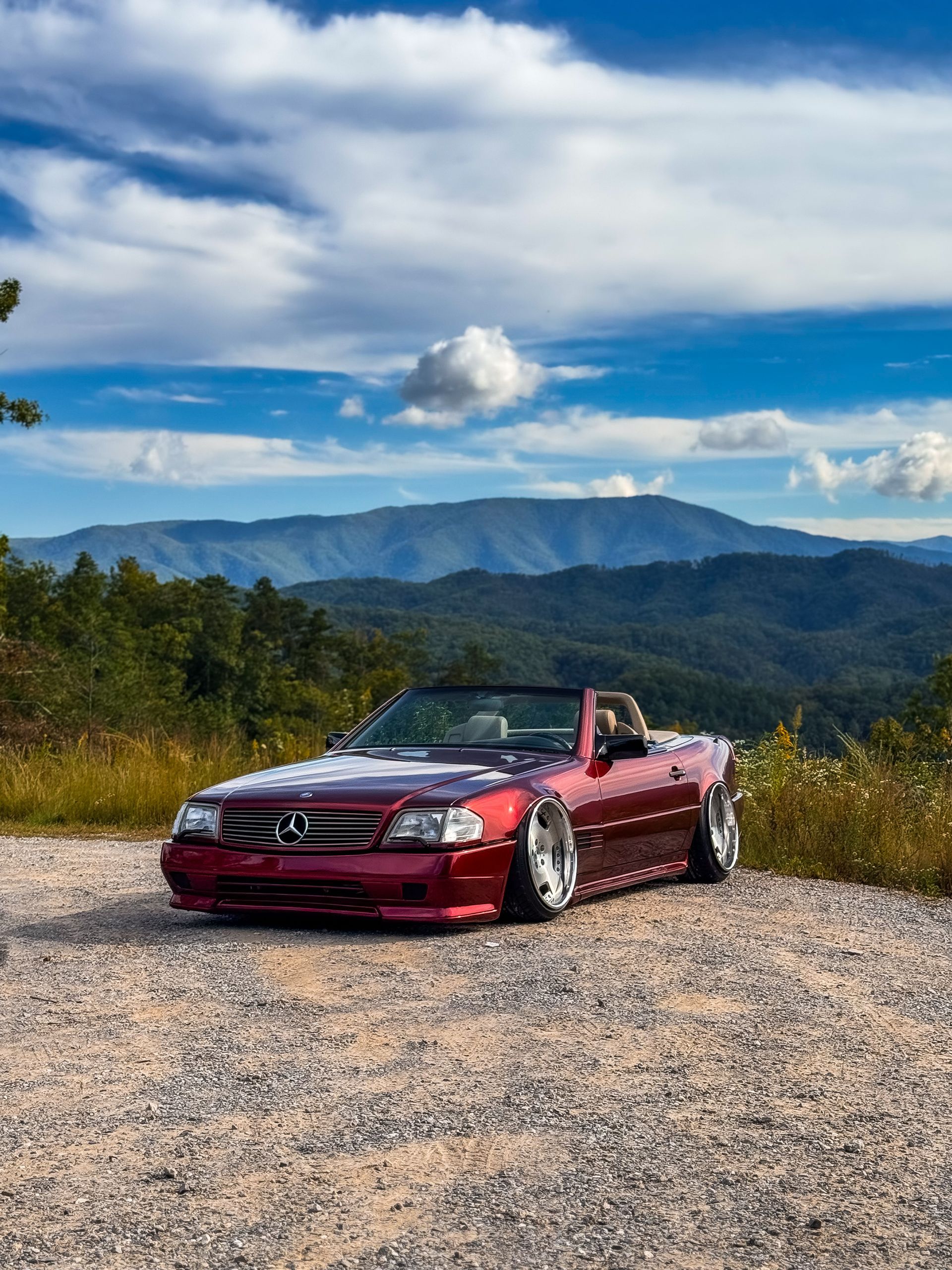 Red convertible car parked on gravel road with mountain backdrop. Cloudy blue sky.