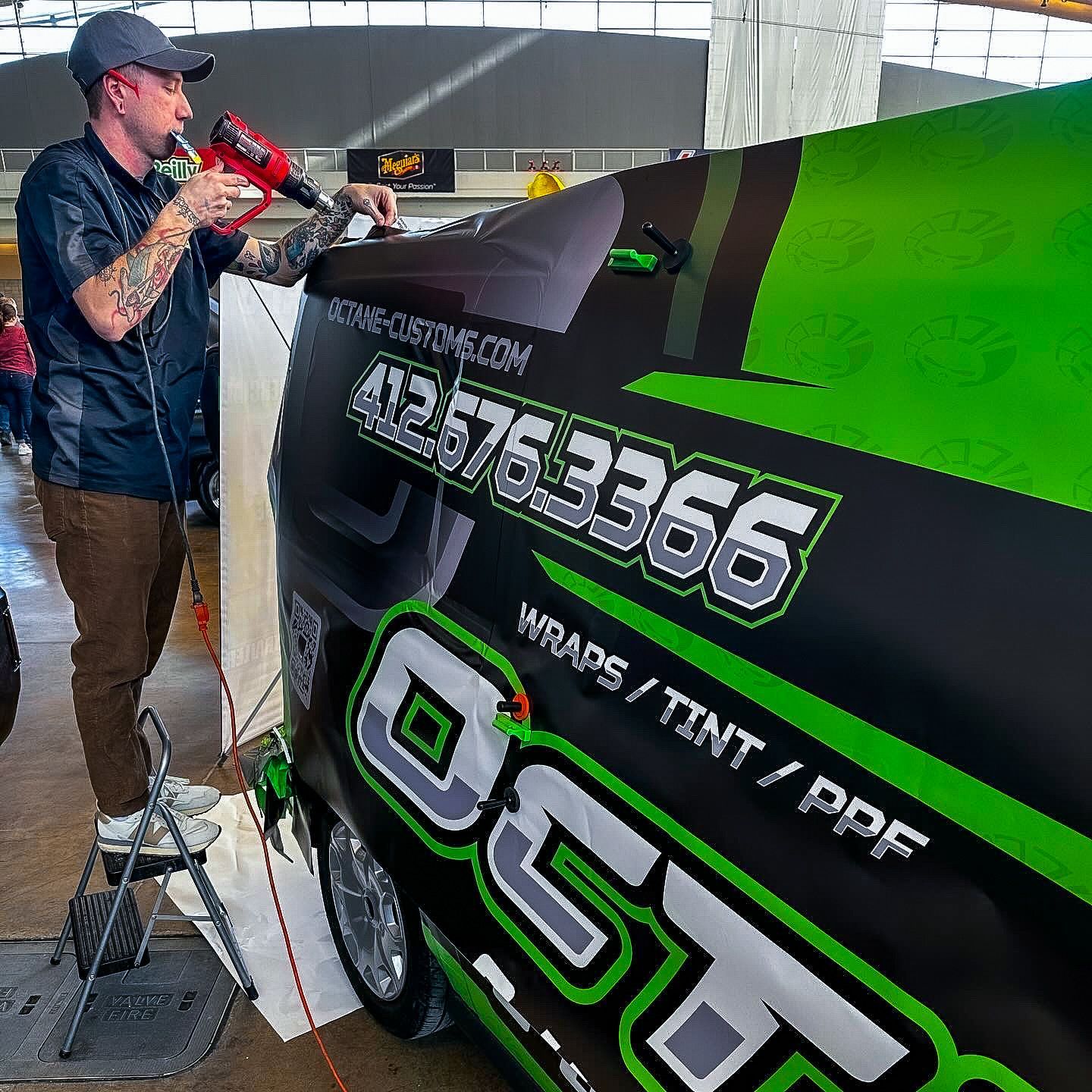 A person applies a vehicle wrap with a heat gun. The van has black and green accents with the business name and contact info on it.