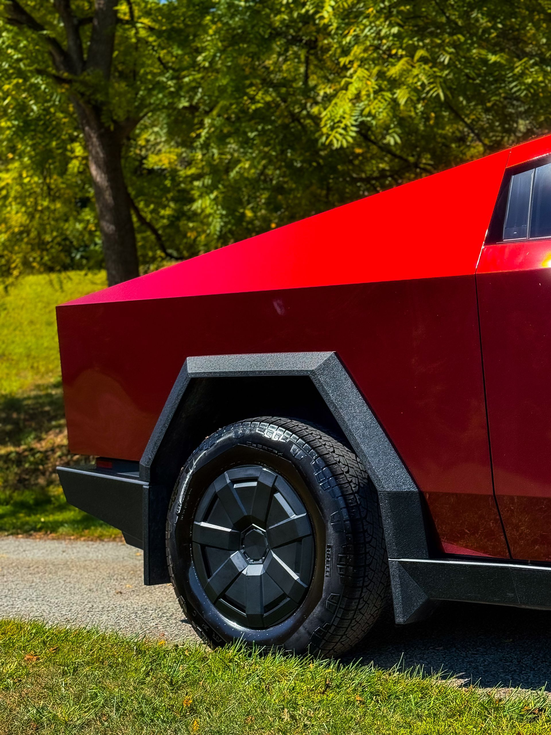 Red Tesla Cybertruck, black wheel, parked on grass. Green trees in background.