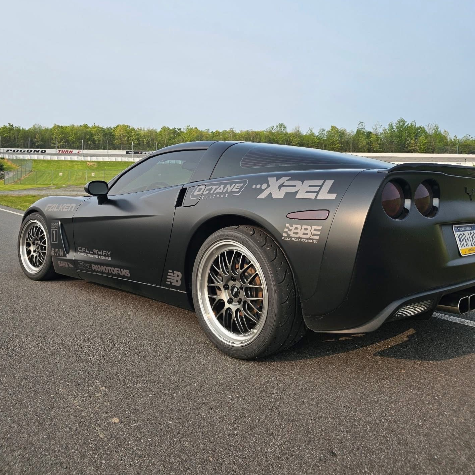Black sports car on a race track, matte finish with sponsor decals, near green trees under a pale sky.