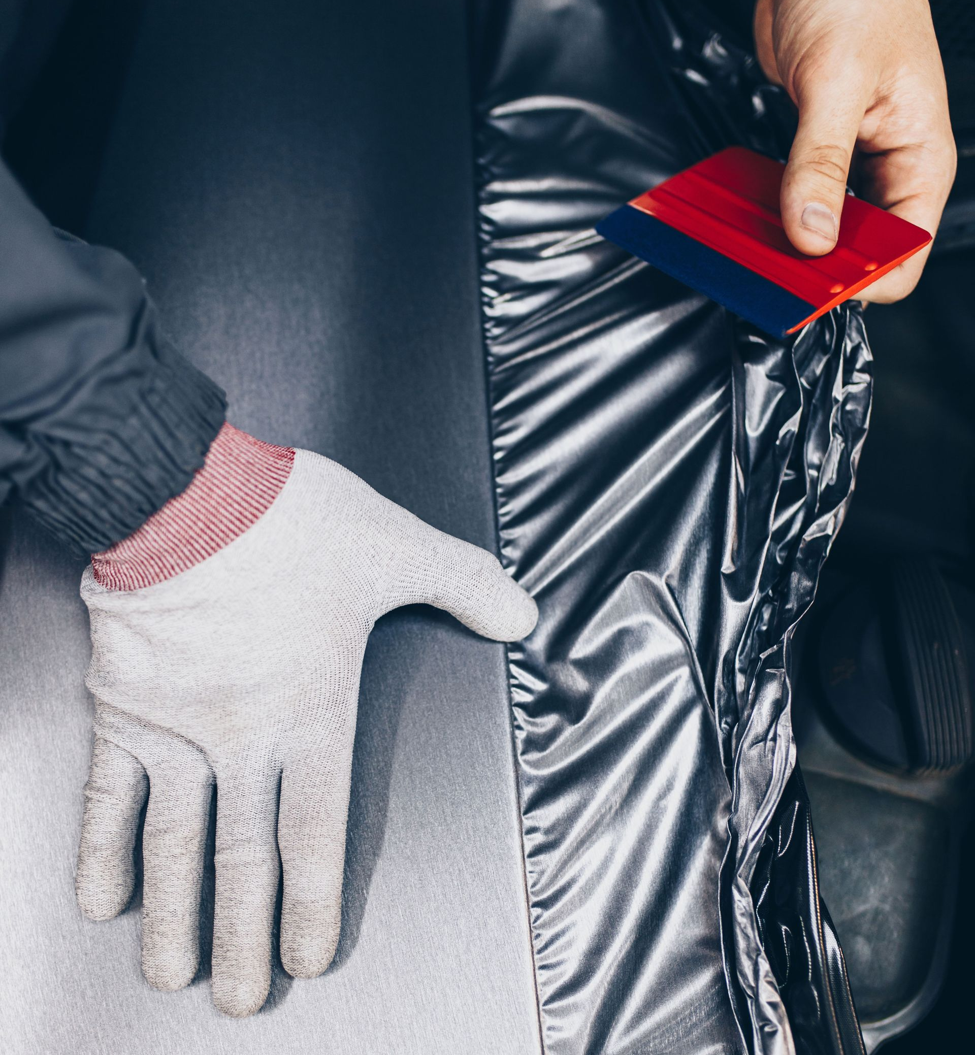Person using a squeegee to apply silver vinyl wrap to a car part.