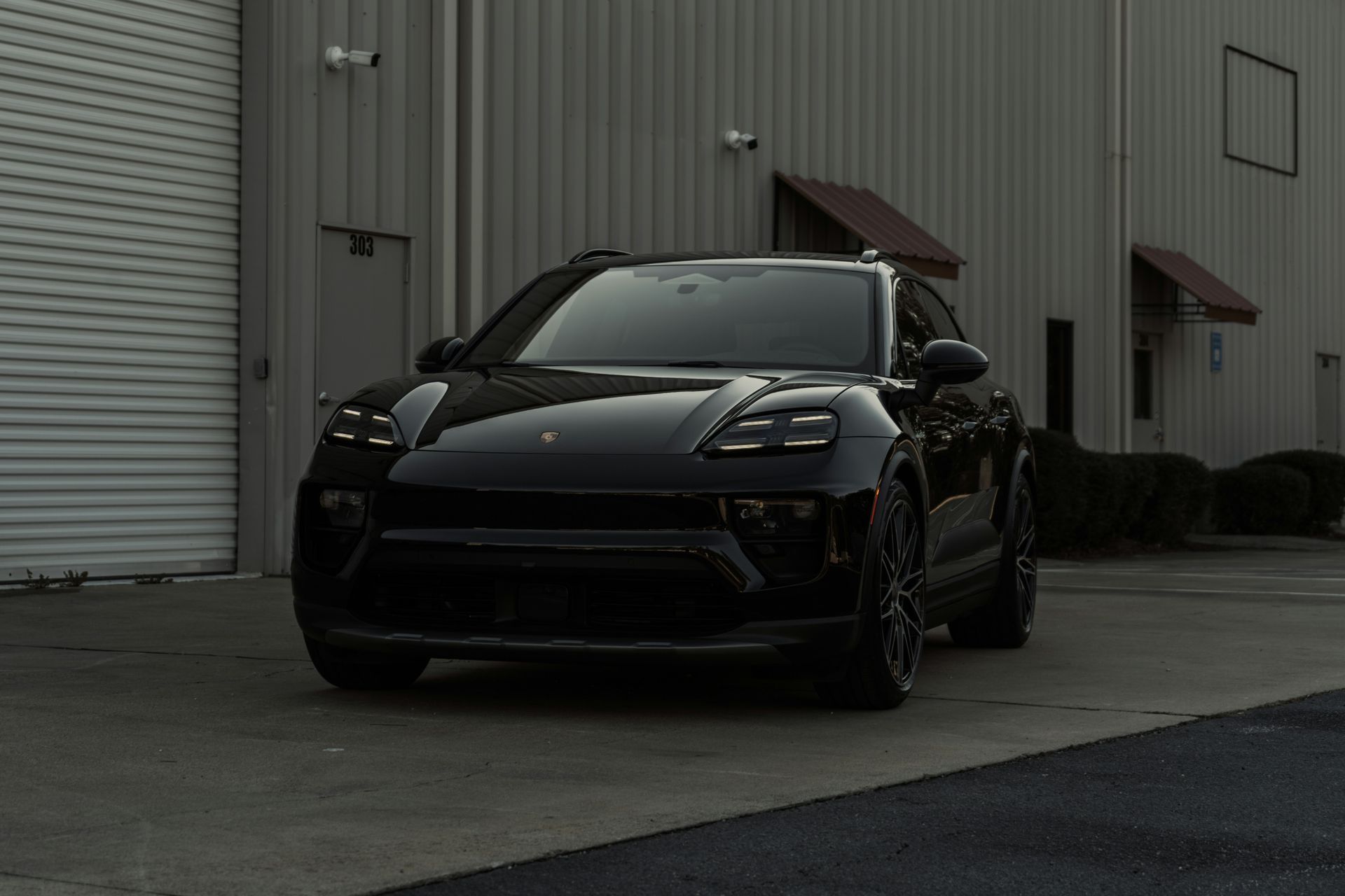 Black Porsche SUV parked in front of a gray building with a roll-up door.