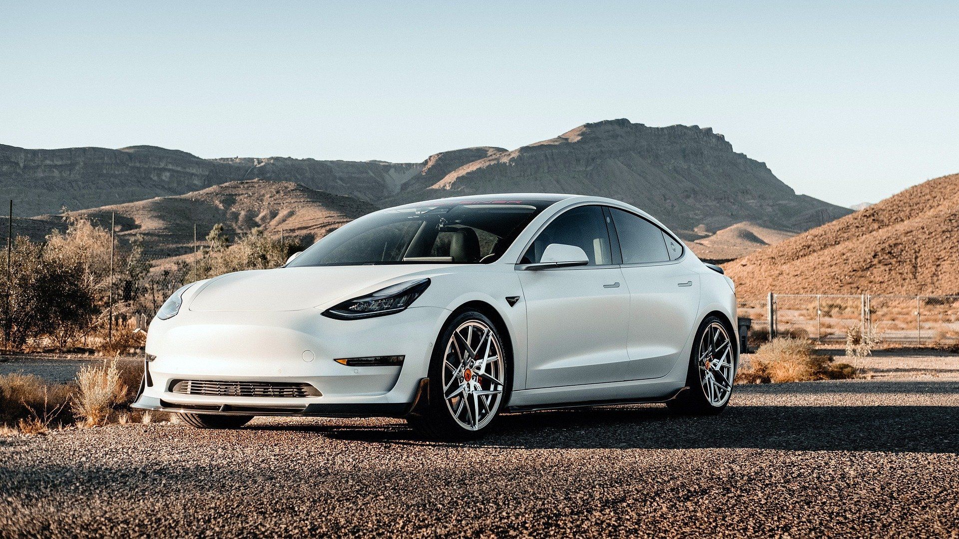White Tesla Model 3 parked on a gravel road, with mountain backdrop under a clear sky.