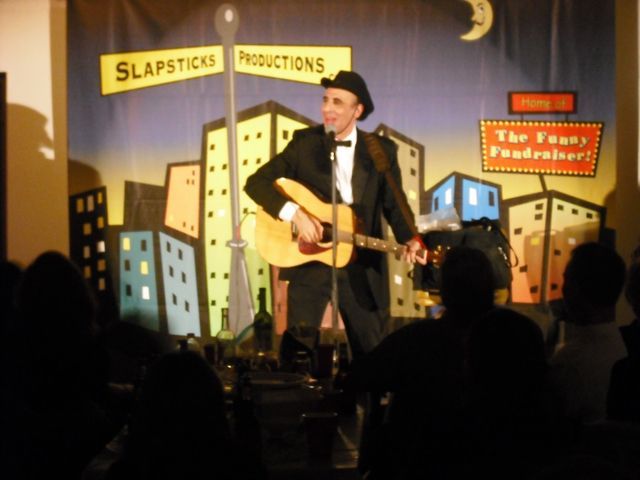 A man singing into a microphone while playing a guitar in front of a slapsticks productions sign