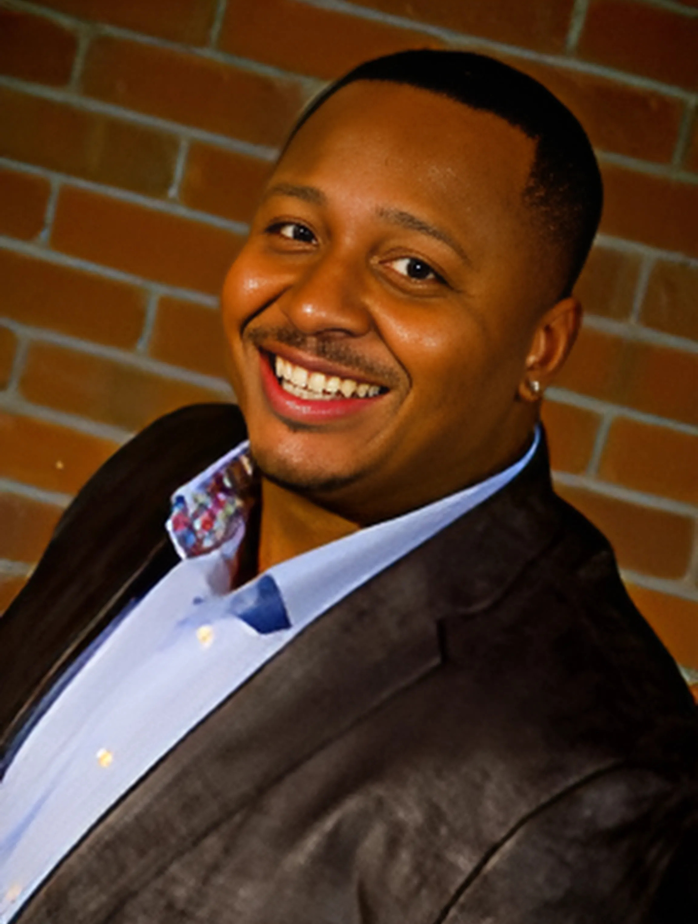 A man in a suit is smiling in front of a brick wall