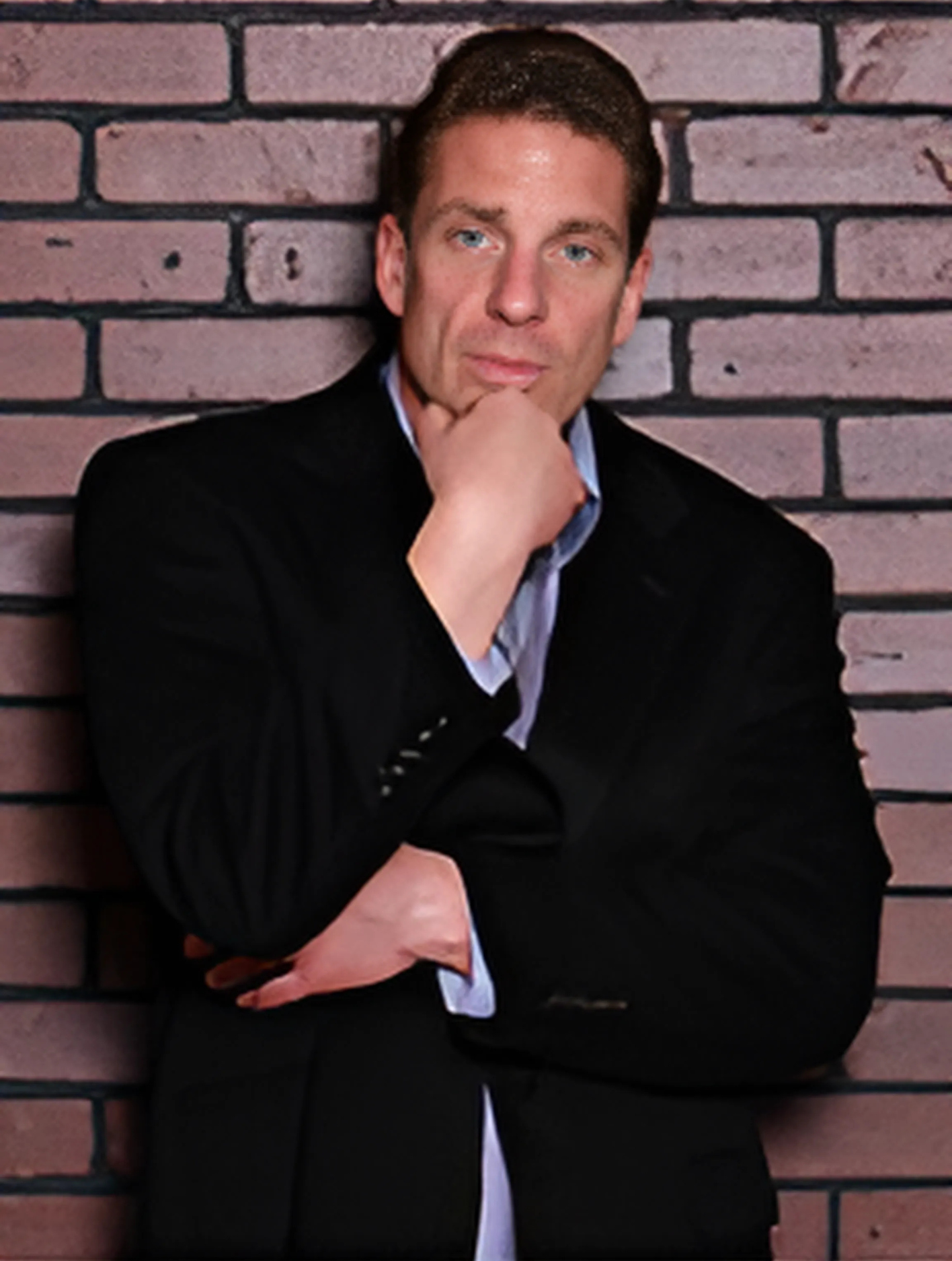 A man in a suit stands in front of a brick wall with his hand on his chin