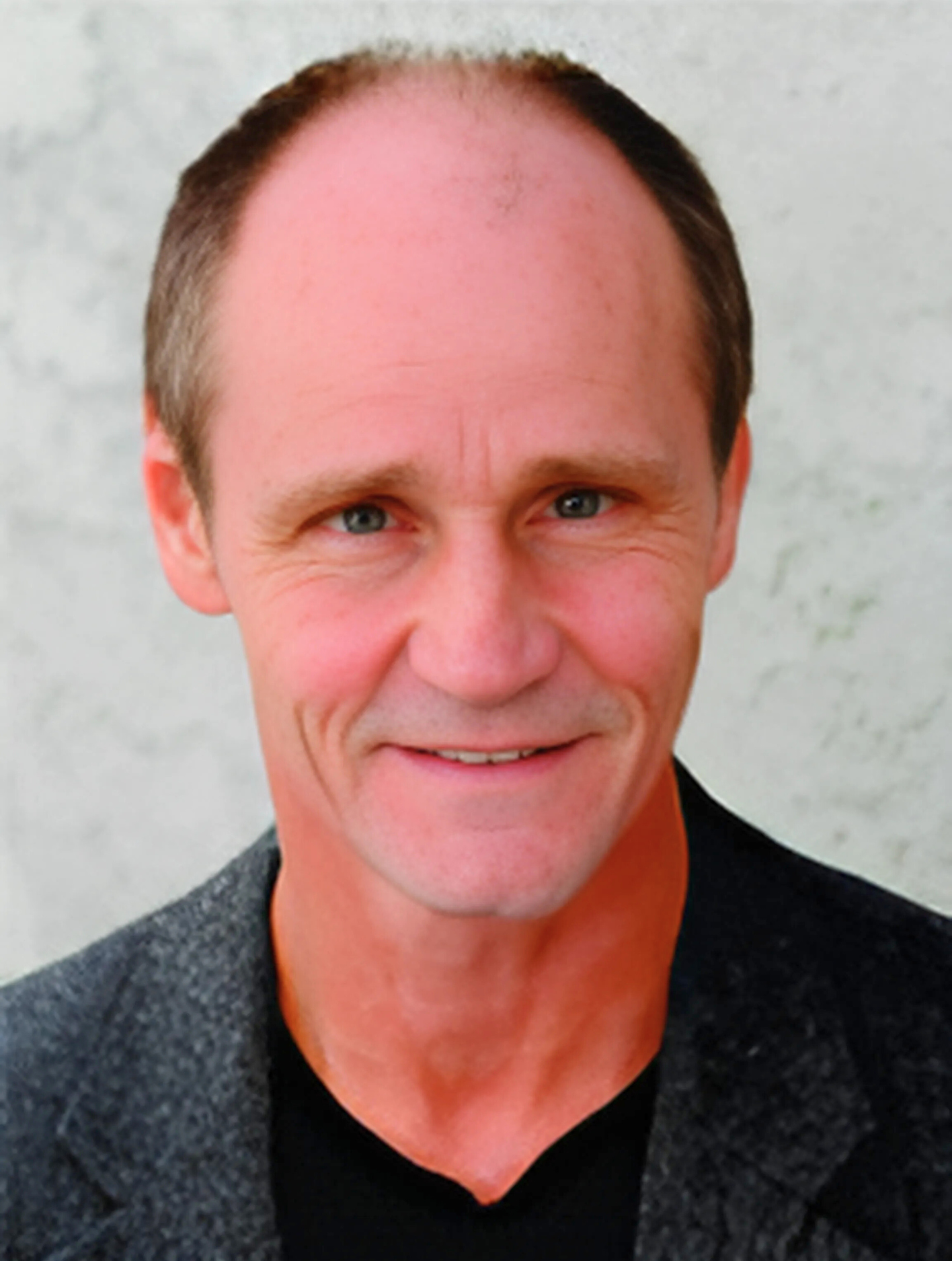 A bald man wearing a black shirt and a grey jacket smiles for the camera
