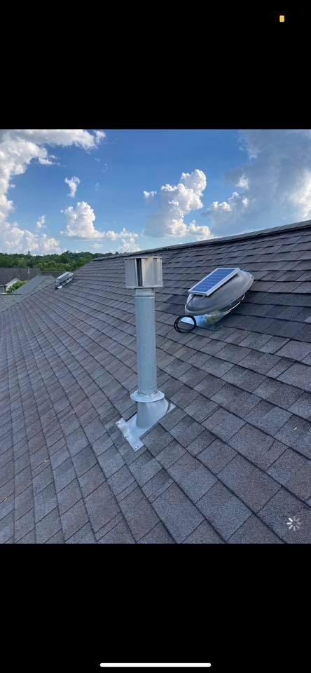 A roof with dark gray shingles has a silver vent pipe and solar-powered vent. Blue sky with clouds.