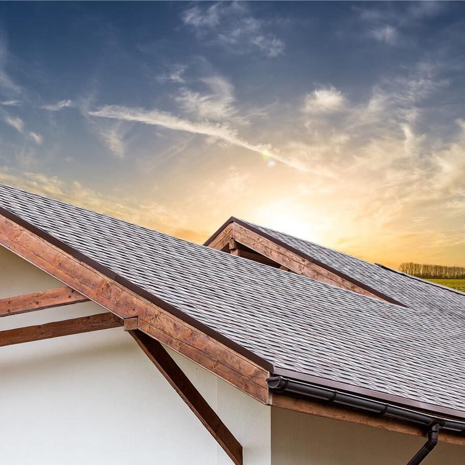Close-up of a house roof with asphalt shingles, brown wood trim, and a sunset sky.