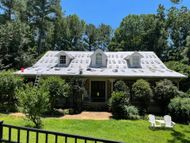 House roof with shingles partially removed, surrounded by trees and greenery. Ladder leans against the side.