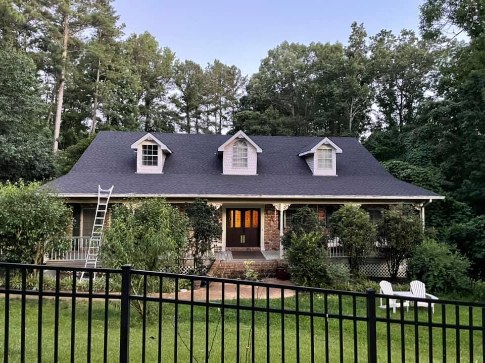 A house with a black roof and three dormers, surrounded by trees and a black fence.