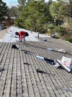 Workers installing roofing shingles on a residential rooftop surrounded by trees on a sunny day.