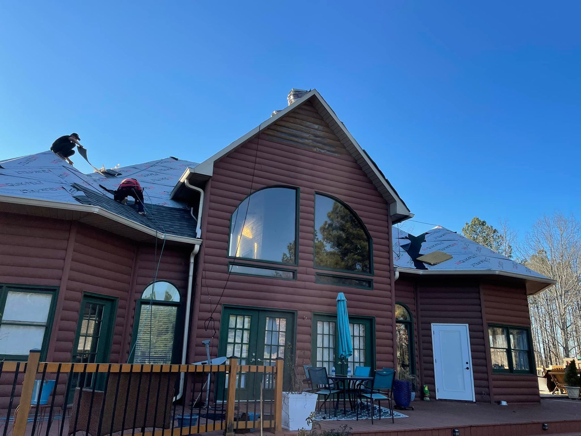 Two people working on a roof of a brown log cabin, sunny day, clear blue sky.