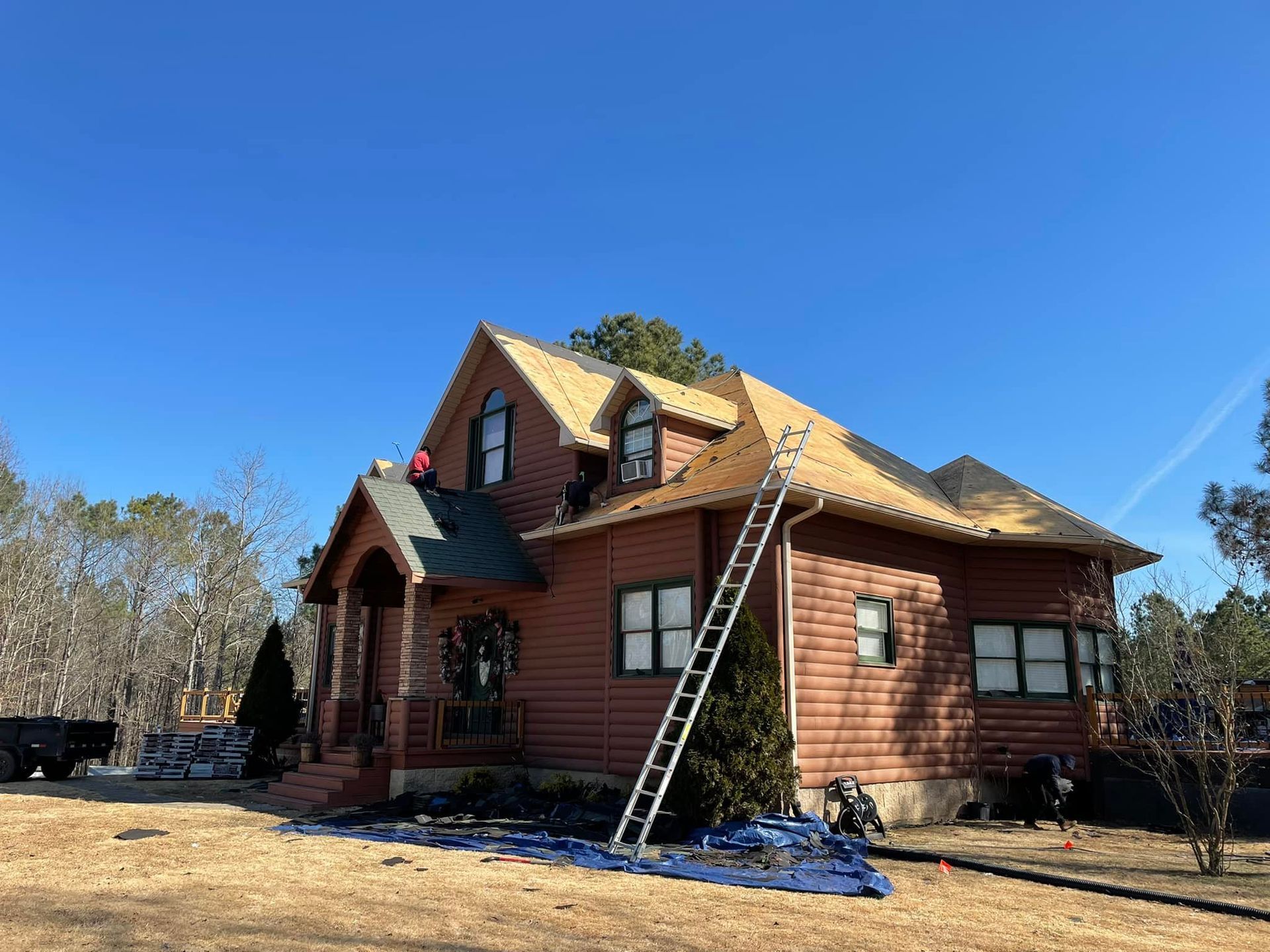House with workers on roof during a roofing project on a sunny day. Ladder and tarp visible.