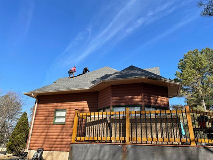 Workers on a roof of a brown wooden house, under a blue sky, with a deck in the foreground.