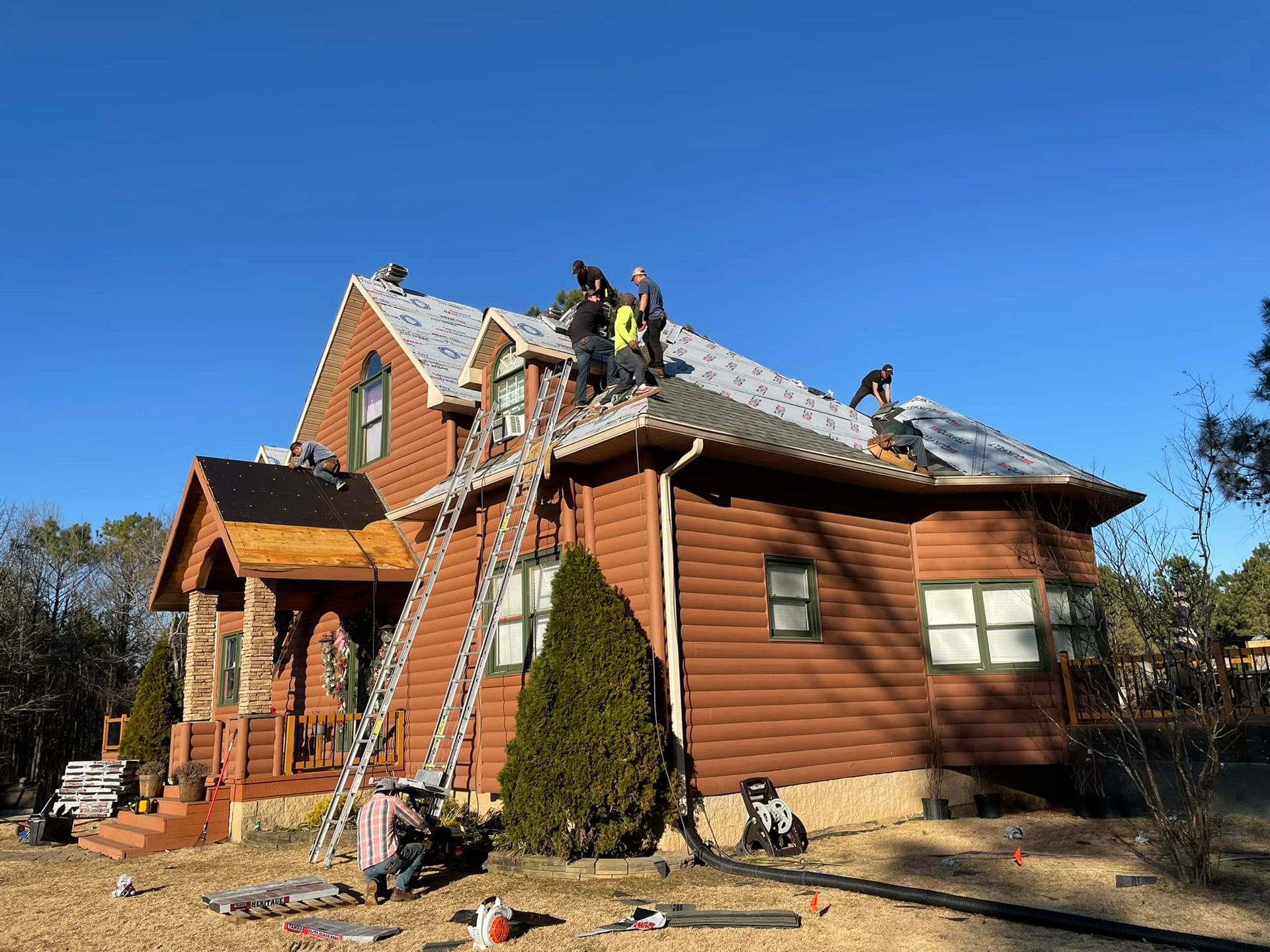 Roofers working on a brown log cabin under a clear blue sky. Ladders propped up.