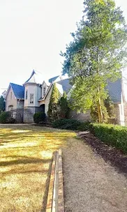 Stone house with a turret and dark blue roof, next to a tree and a gravel path.