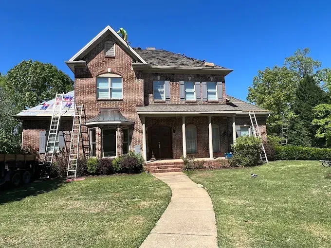 Brick house with roof under construction; ladders, blue sky, green lawn.