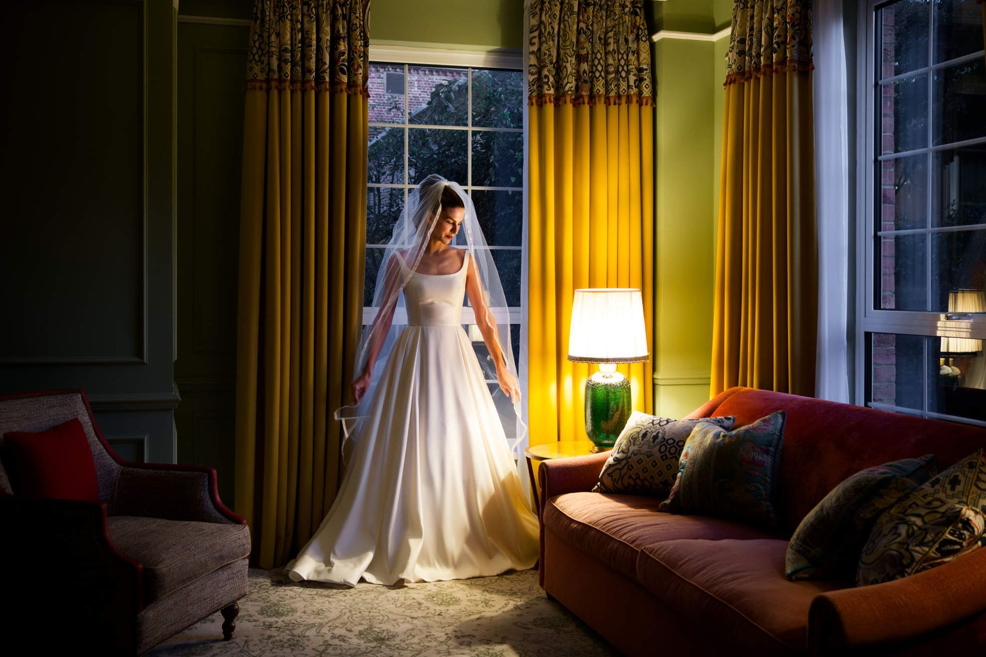 A woman in a wedding dress is standing in front of a window in a living room.