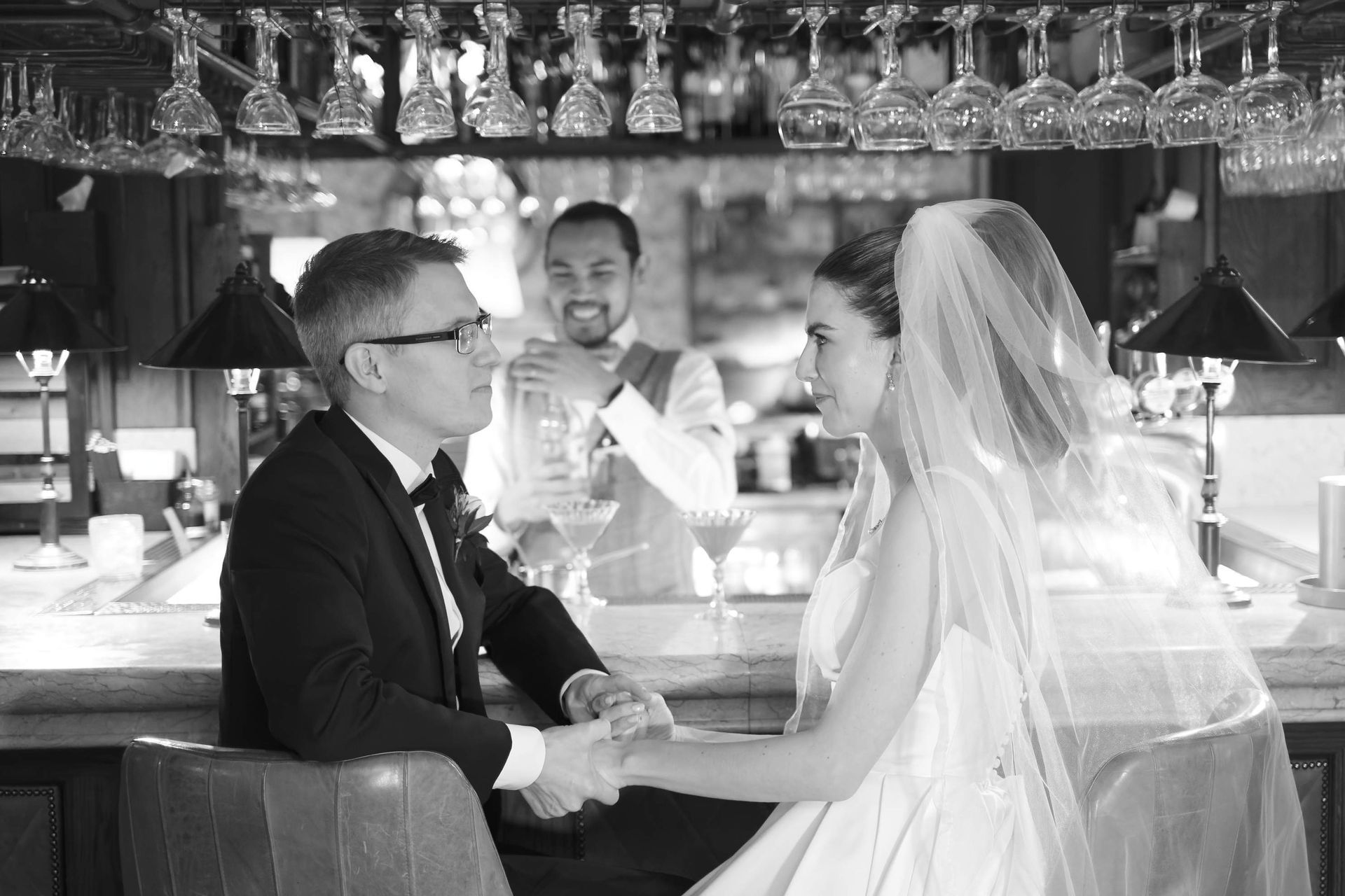 A bride and groom are sitting at a bar holding hands.