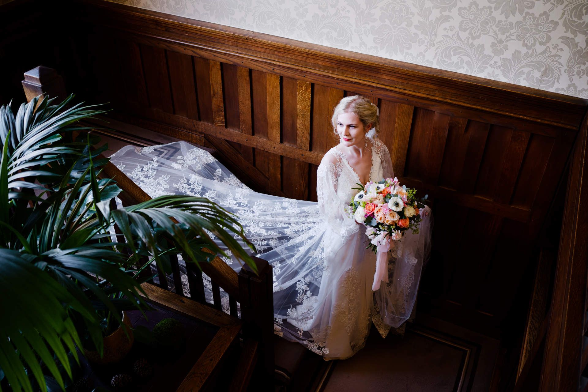 A bride in a long wedding dress is sitting on a staircase holding a bouquet of flowers.