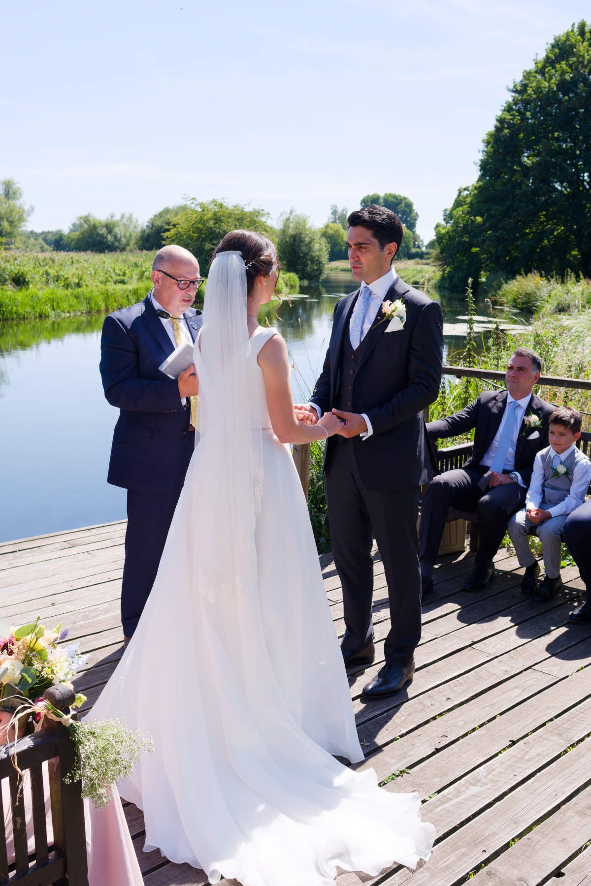 A bride and groom are holding hands during their wedding ceremony on a dock.