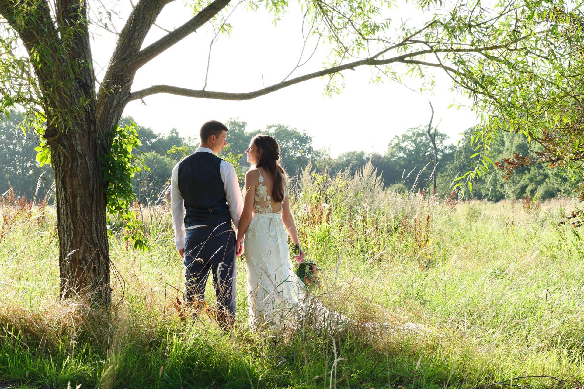 A bride and groom are standing under a tree in a field holding hands.