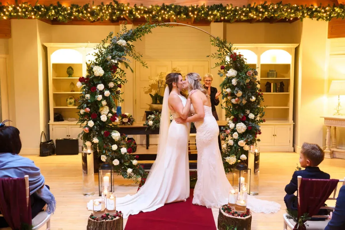 gay couple are kissing during a wedding ceremony in front of a floral arch.