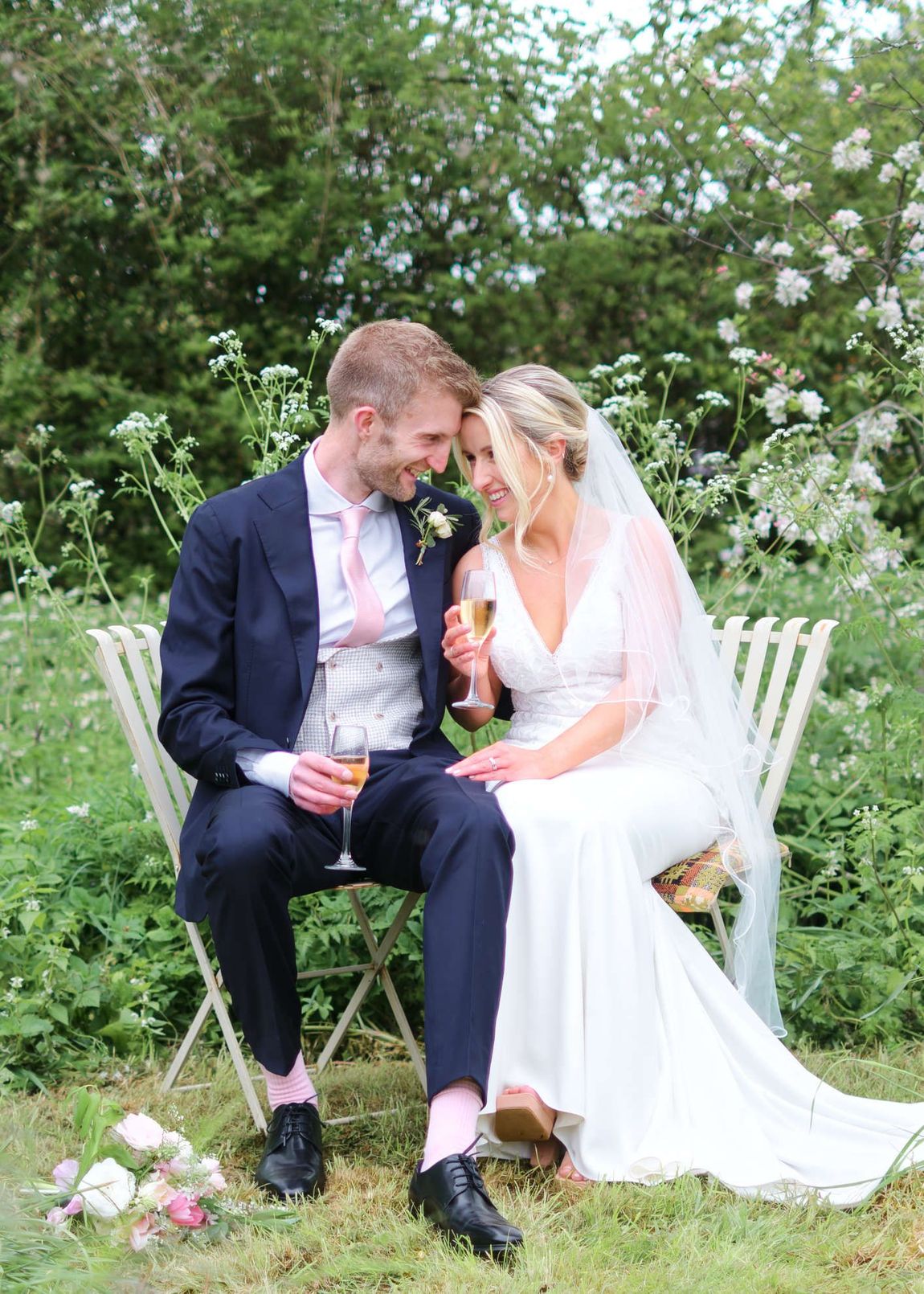 A bride and groom are sitting on a bench in the grass drinking champagne.