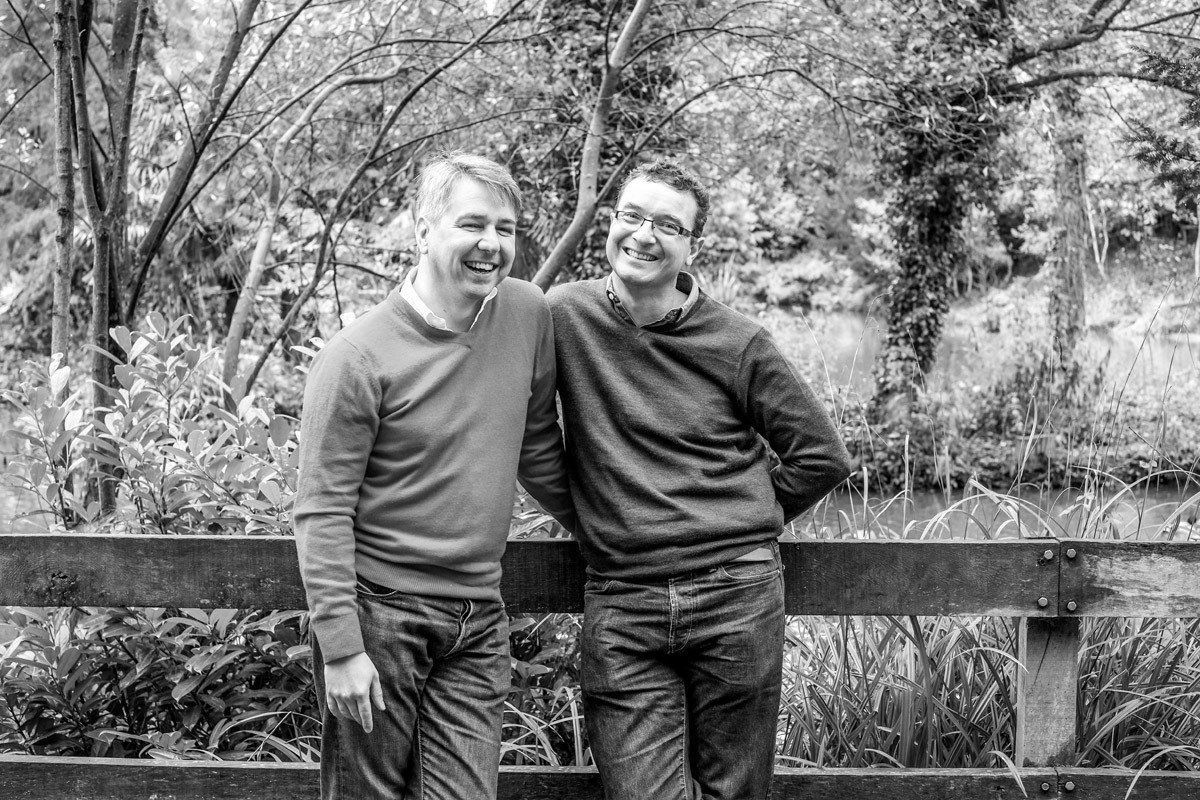 Two men are standing next to each other in front of a wooden fence.