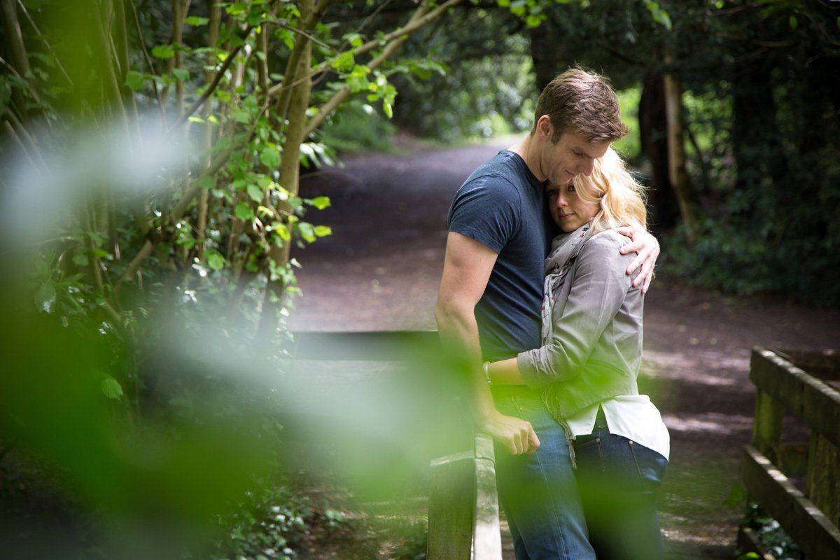A man and a woman are hugging on a wooden bridge in the woods.