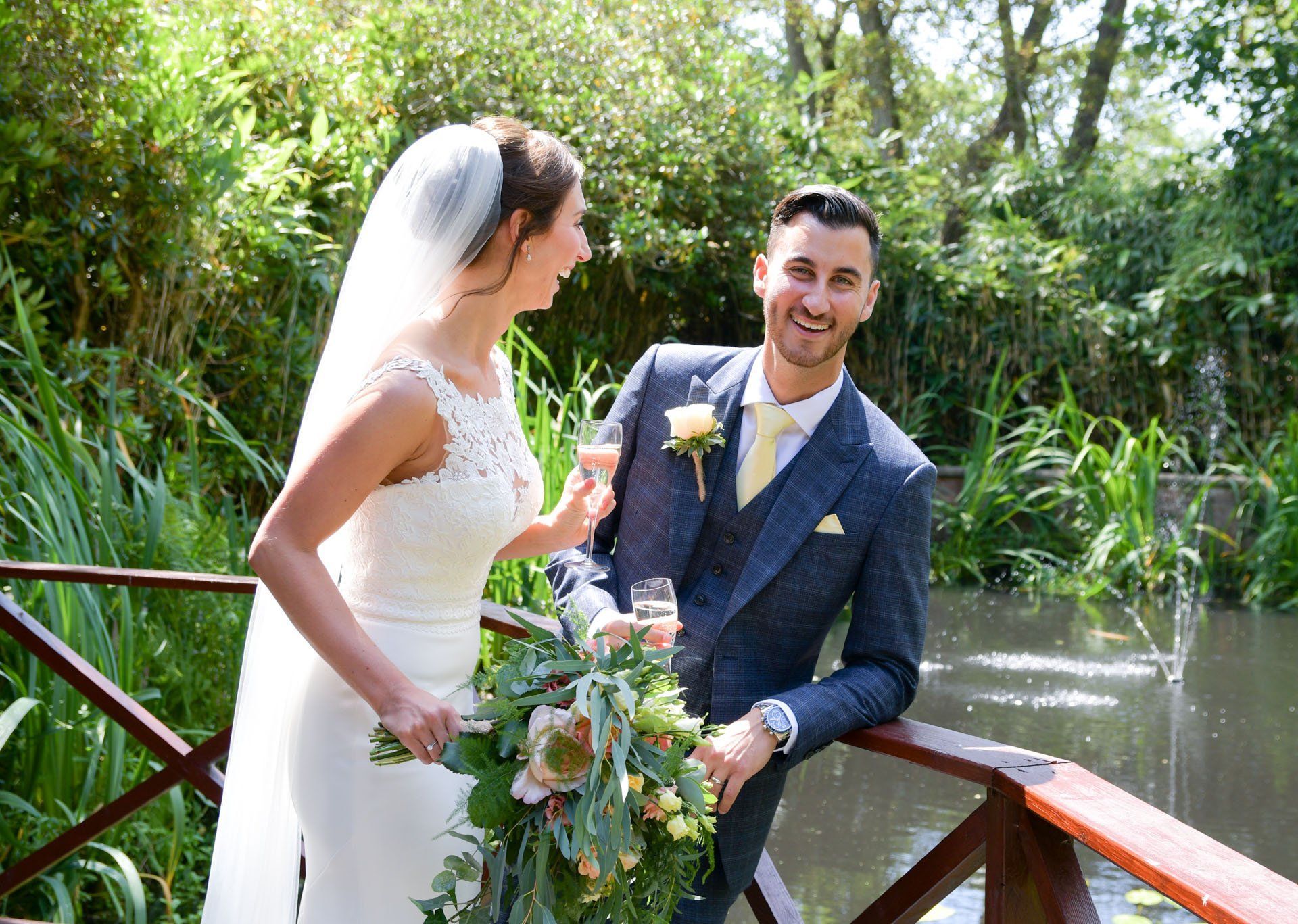 A bride and groom are standing on a bridge holding champagne glasses.