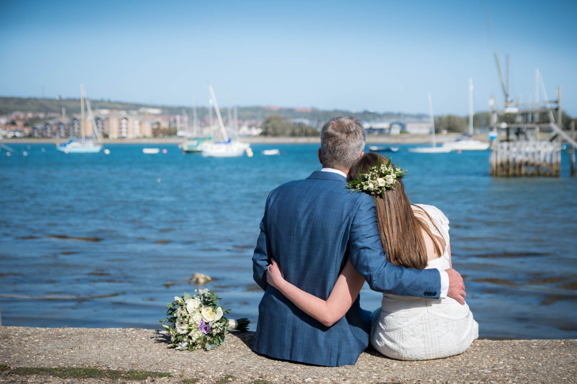 A bride and groom are sitting on a wall overlooking the water.