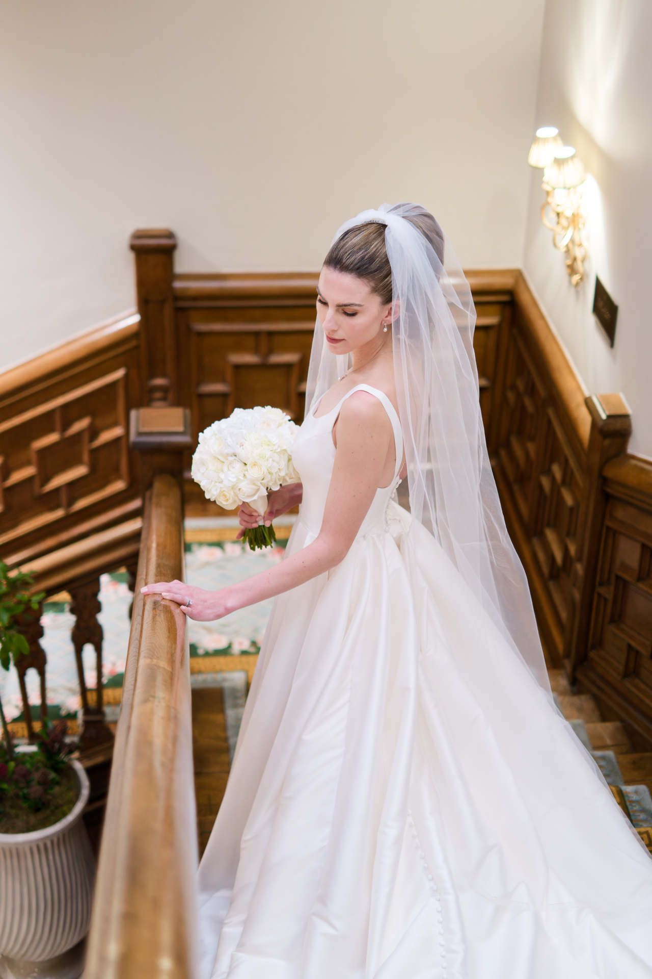 A bride in a wedding dress and veil is standing on a staircase holding a bouquet of flowers.