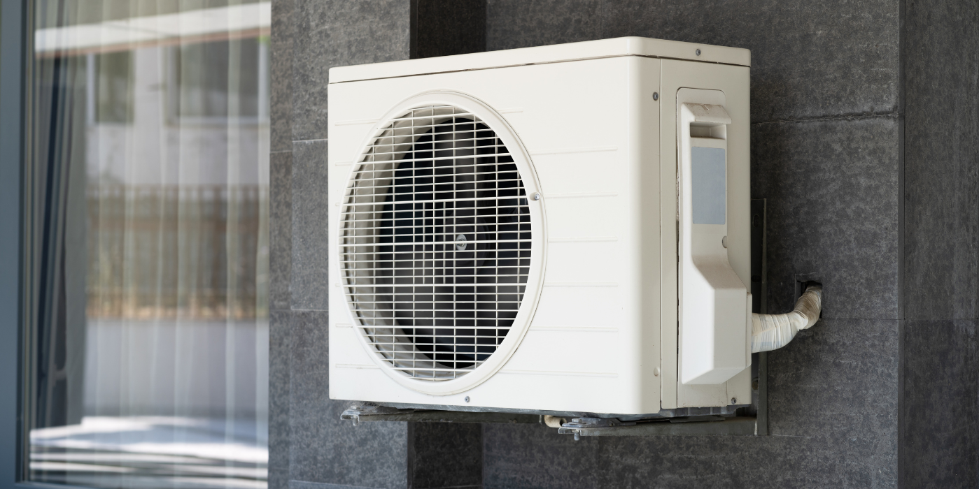 A white air conditioner is mounted on the side of a building next to a window.